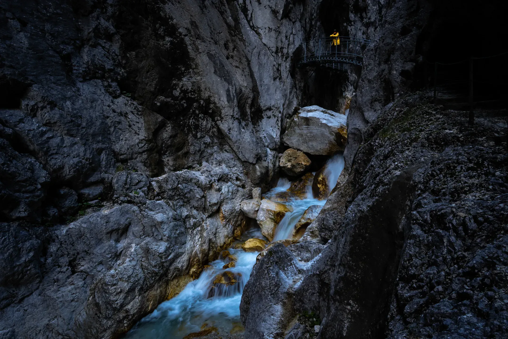 Wildbach rauscht durch eine dunkle Felsklamm, oben steht eine Person in gelber Jacke auf einer Metallbrücke