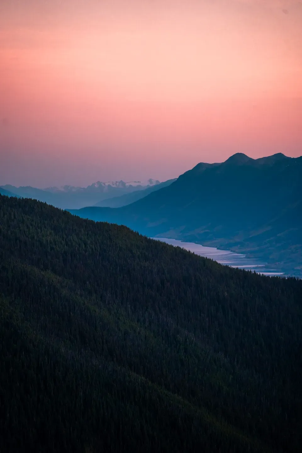 Bewaldeter Berghang im Vordergrund, dahinter ein Fluss im Tal und Bergsilhouetten unter rosa Abendhimmel