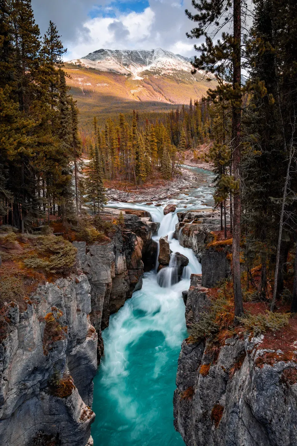 Türkiser Fluss stürzt als Wasserfall in eine enge Felsschlucht, dahinter Nadelwald und ein schneebedeckter Berg