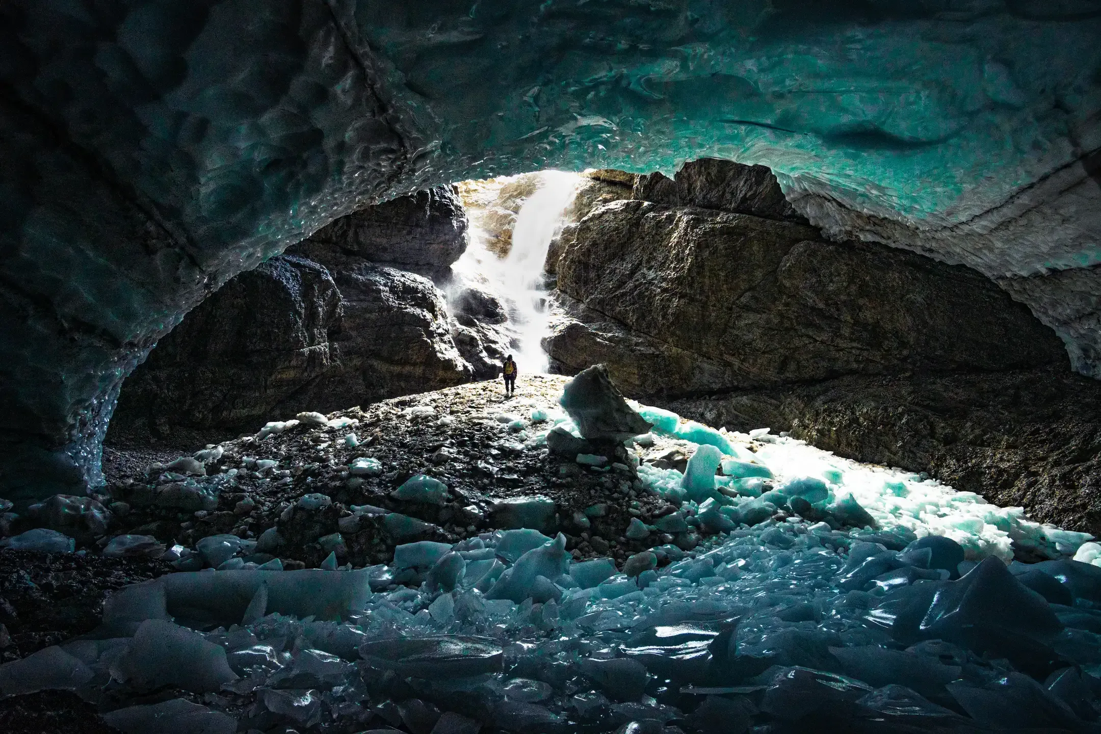 Blick aus einer Eishöhle auf einen Wasserfall, eine Person steht klein im Licht, vorne türkise Eisbrocken