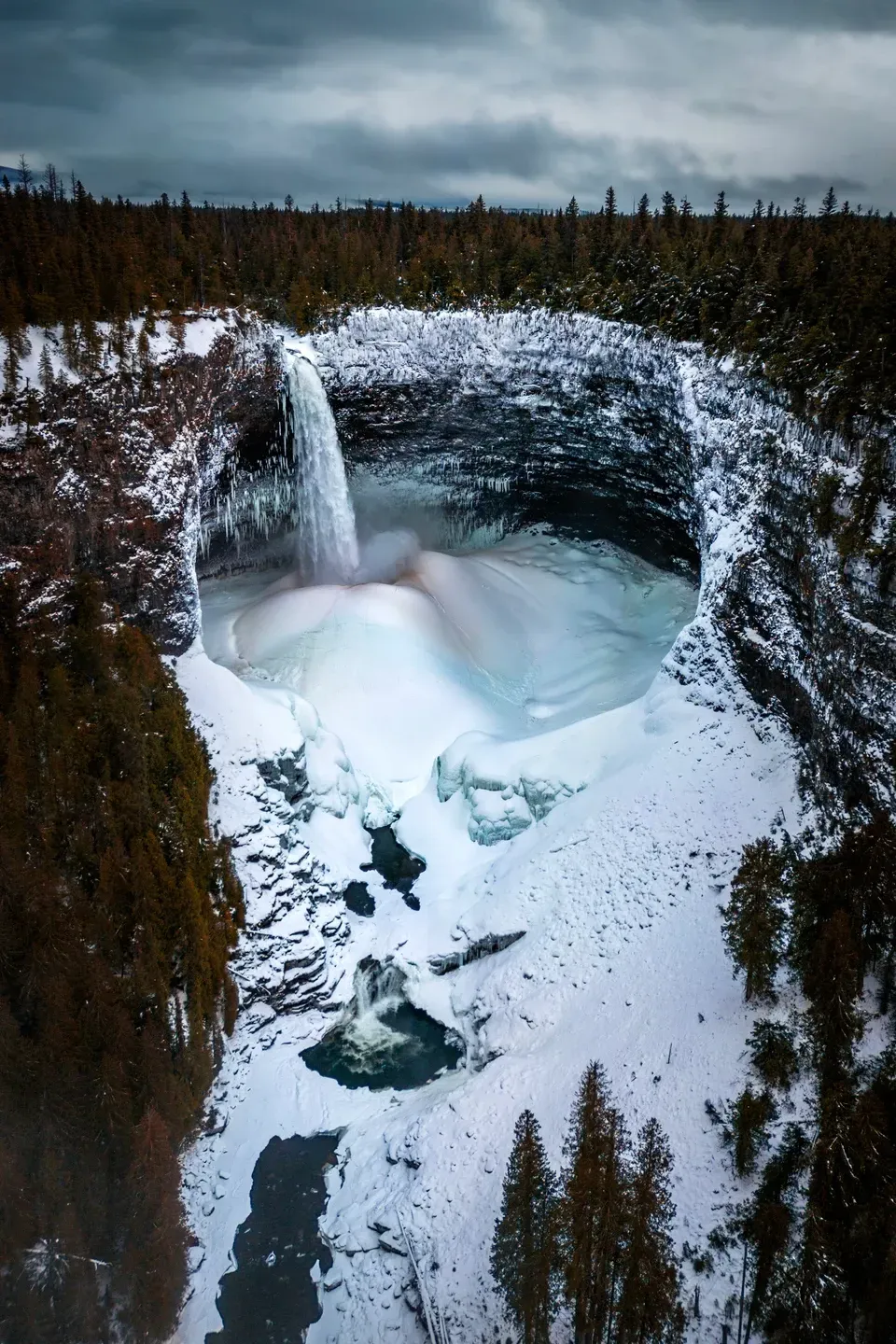 Gefrorener Wasserfall stürzt in einen verschneiten, runden Felskessel, umgeben von Nadelwald unter grauem Himmel