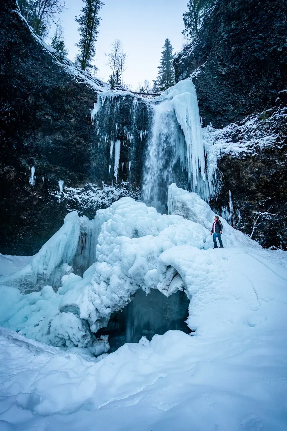 Person in roter Jacke steht auf einem Eishügel vor einem gefrorenen Wasserfall mit langen Eiszapfen