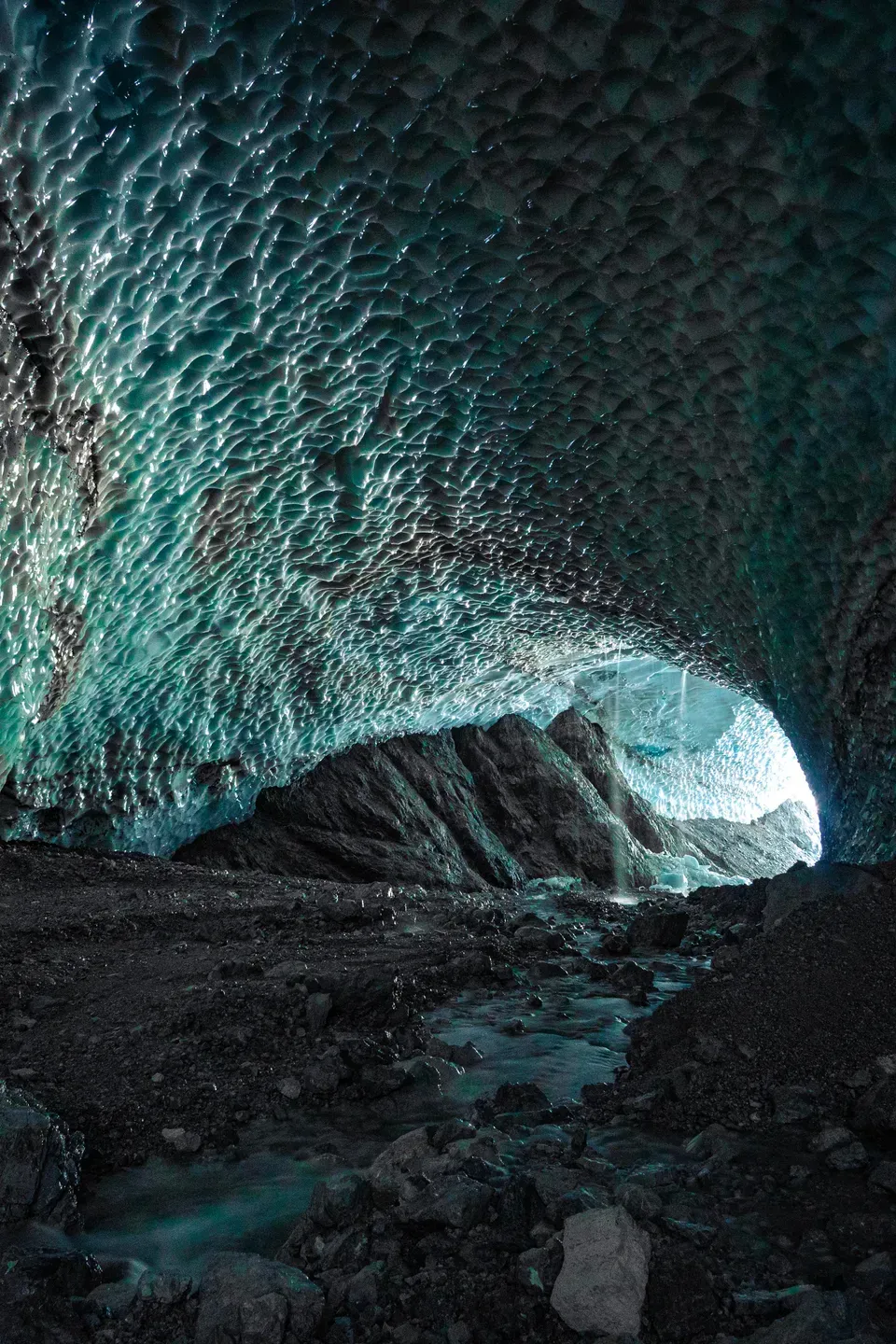 Gewölbe einer Eishöhle mit türkis schimmernder, gemusterter Decke, ein Bach fließt über Geröll zum Ausgang