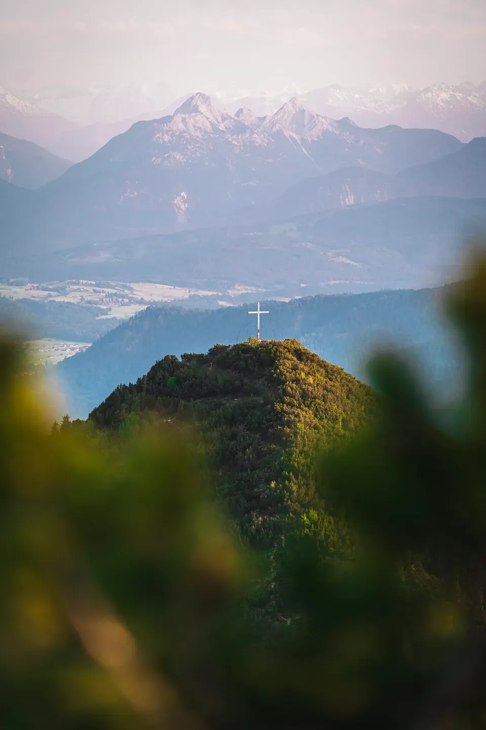 Bewaldeter Berggipfel mit Gipfelkreuz im Abendlicht, dahinter Täler und Alpengipfel im Dunst