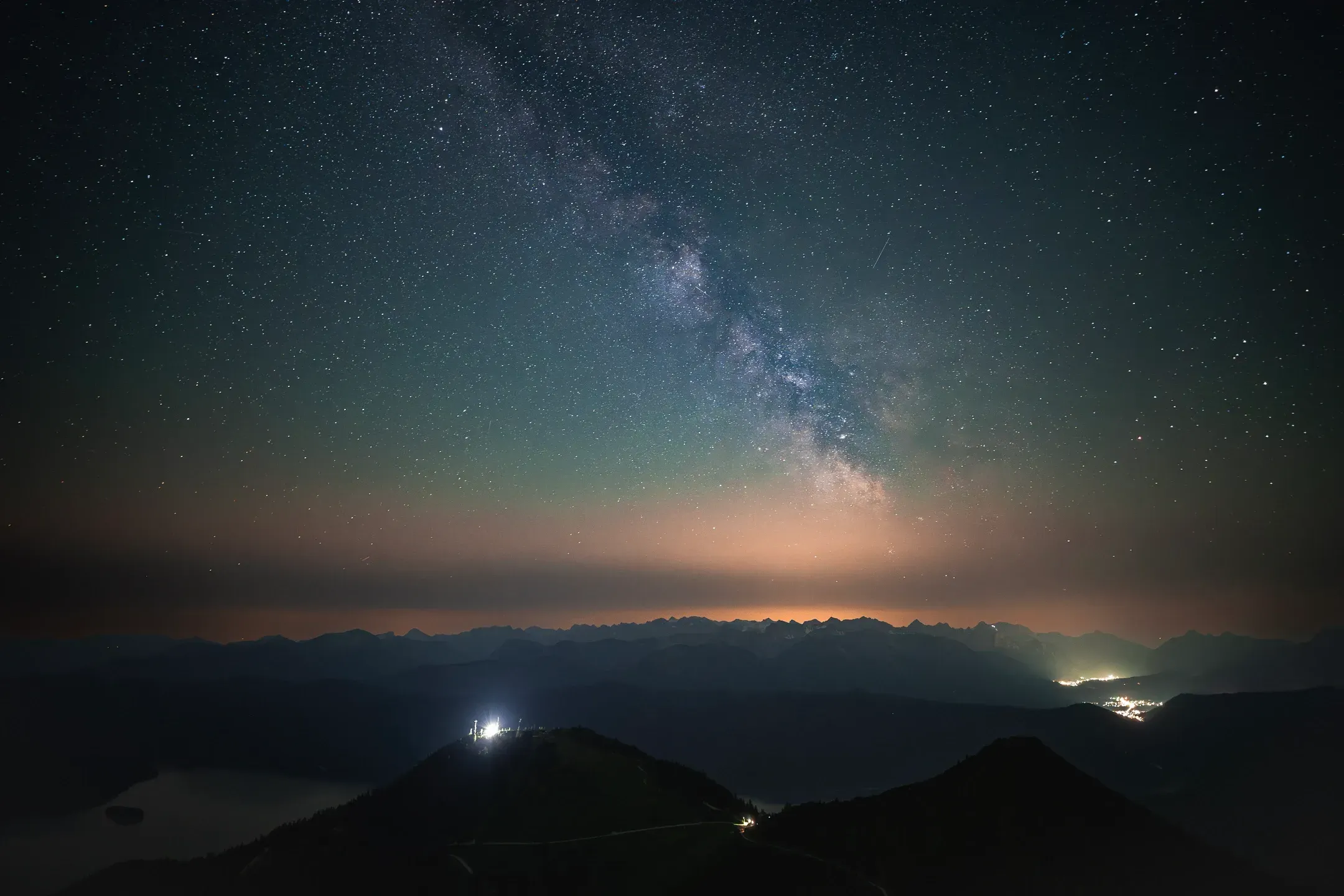 Milchstraße über nächtlichen Bergketten, auf einem Gipfel leuchten Lichter und am Horizont glüht ein Ort