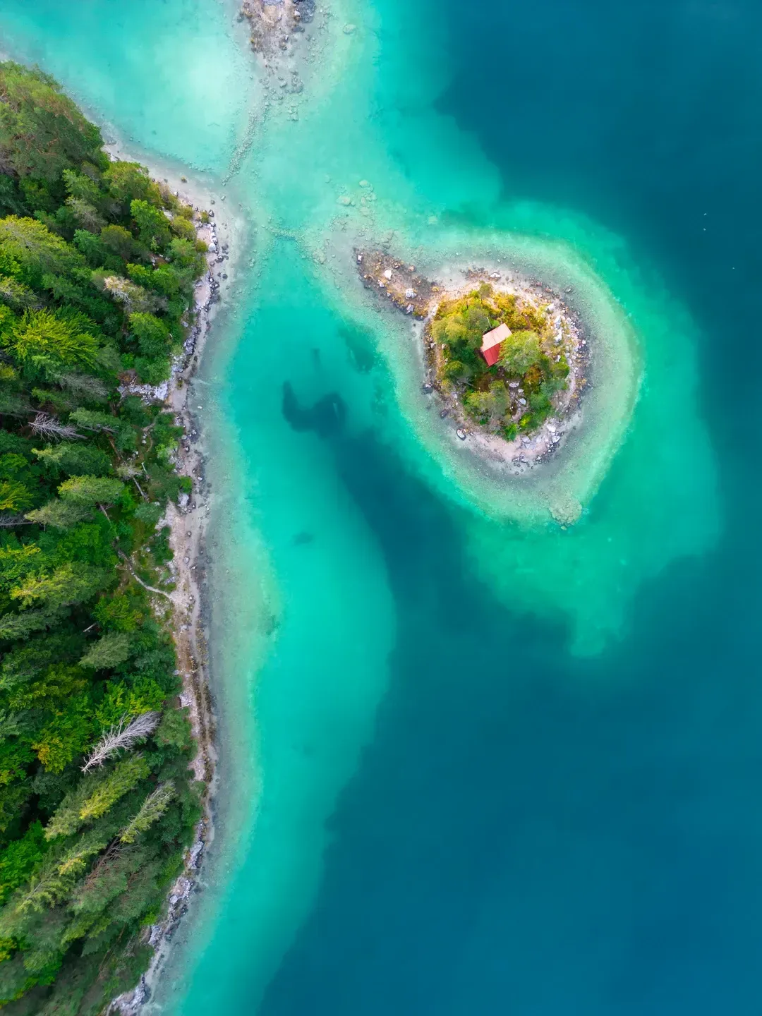 Luftaufnahme einer kleinen Insel mit roter Hütte in einem türkisen Bergsee neben bewaldetem Ufer