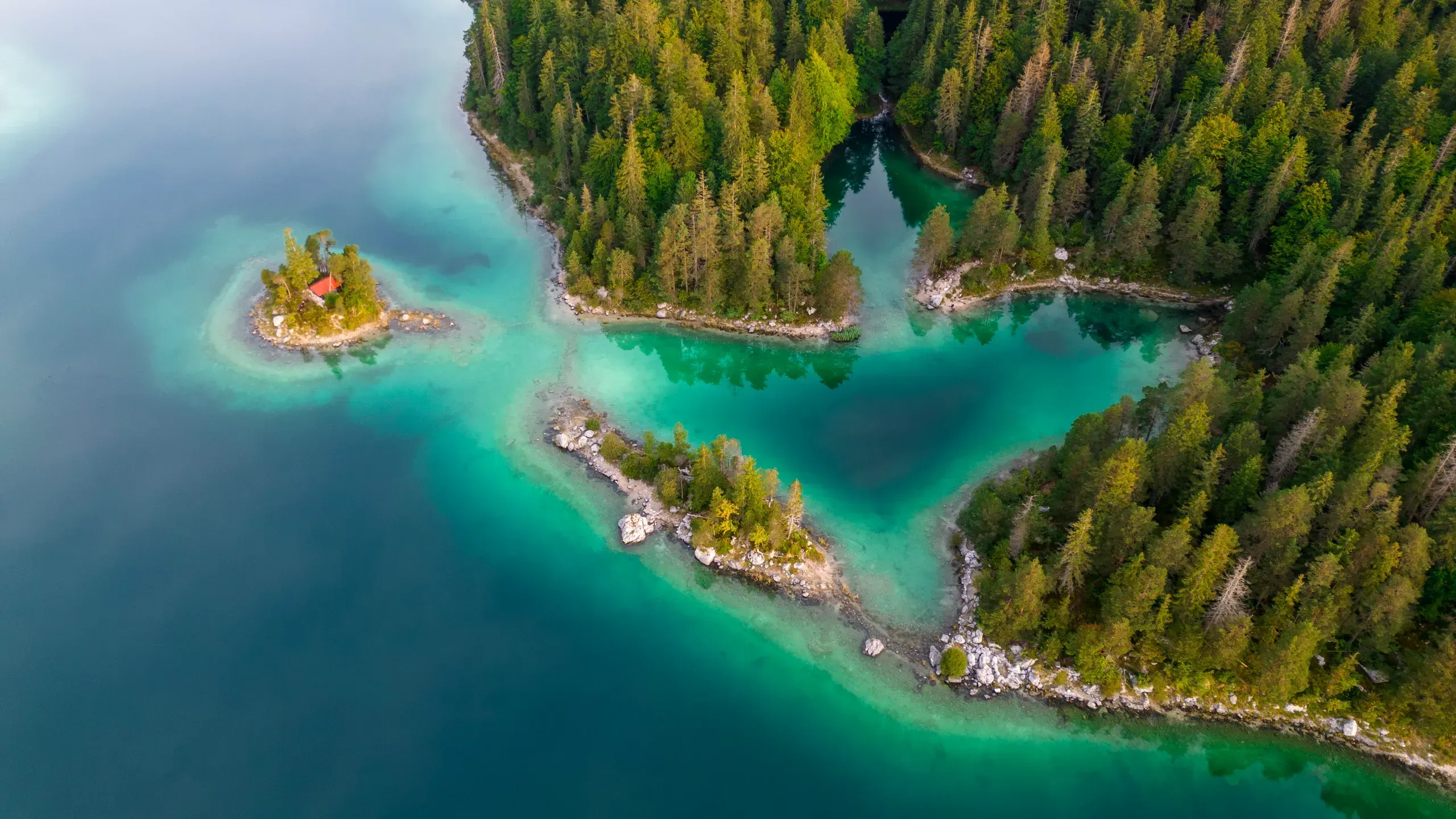 Drohnenblick auf einen türkisen Bergsee mit bewaldeten Buchten, Landzungen und einer kleinen Insel mit roter Hütte